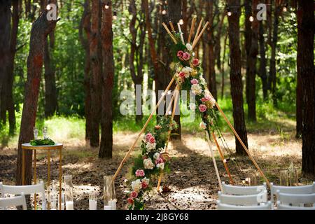 Wunderschöne böhmische Tipi-Bogendekoration auf der Hochzeitsfeier im Freien im Pinienwald mit Kegeln. Stühle, floristische Blumenkompositionen aus Rosen Stockfoto