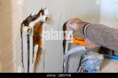 Hände von Klempnern in Handschuhen schneiden ein Kunststoffrohr mit einem Rohrschneider. Installation von Sanitär im Raum. Stockfoto