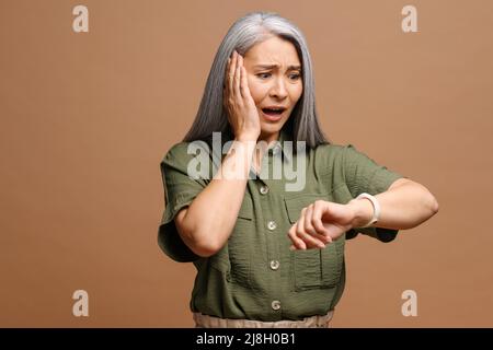 Porträt einer schockierten, reifen Frau, die Hand mit Armbanduhr hält und sie auf einem braunen Hintergrund mit einem überraschenden Ausdruck betrachtet Stockfoto