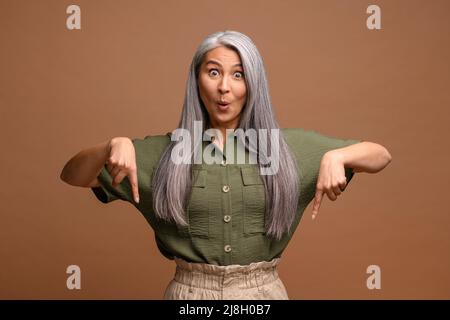 Fröhliche positive Frau, die mit den Fingern nach unten zeigt, um Platz für Ihre Werbung zu schaffen, und mit einem überraschten Gesicht auf die Kamera blickt. Innenaufnahme des Studios isoliert auf braunem Hintergrund Stockfoto