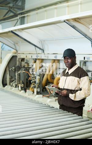 Afrikanischer ernsthafter Arbeiter in Uniform, der während seiner Arbeit mit der Maschine in der Fabrik digitale Tablets verwendet Stockfoto