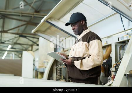 Afrikanischer Arbeiter in Uniform mit digitalem Tablet, um die Maschine in der Fabrik zu steuern Stockfoto