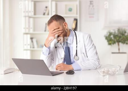 Gestresster männlicher Arzt, der in einem Büro mit einem Laptop sitzt und seinen Kopf hält Stockfoto