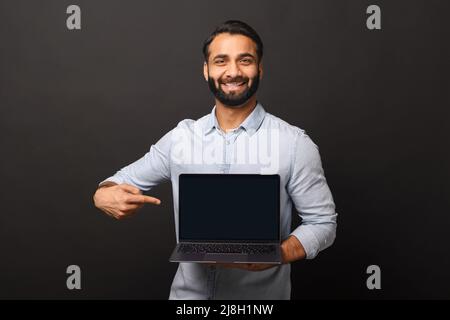 Glücklicher indischer Mann, der einen Laptop mit leerem Bildschirm hält und mit dem Finger auf ihn zeigt, hispanic-Mitarbeiter, der neue Computer-App, Werbung und Werbekonzept vorstellt Stockfoto