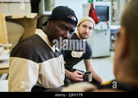 Reifer afrikanischer Mann in Uniform im Gespräch mit seinen Kollegen während der Kaffeepause im Lager Stockfoto