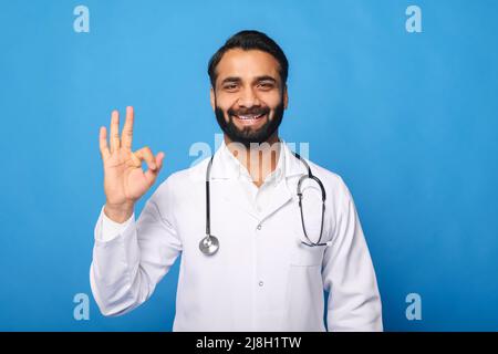 Freundlicher, fröhlicher indischer Arzt in weißem Kleid und Stethoskop, der mit Hand und Fingern ein Schild mit OK zeigt, lächelnd auf die Kamera blickt, Mann in Ärzteuniform isoliert auf blau Stockfoto