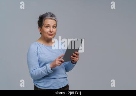 Wunderbare reife Frau 50s grauhaarig mit digitalem Tablet, das in sozialen Medien arbeitet oder überprüft. Hübsche Frau in blauer Bluse isoliert auf weiß. Ältere Menschen und Technologien. Getöntes Bild. Stockfoto