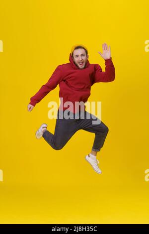 Lustiges Porträt eines jungen Mannes in Freude springen hoch tragen lässigen roten Hoodie und Jeans isoliert auf gelbem Hintergrund. Humorvoller Hipster-Typ im Sprung. Stockfoto