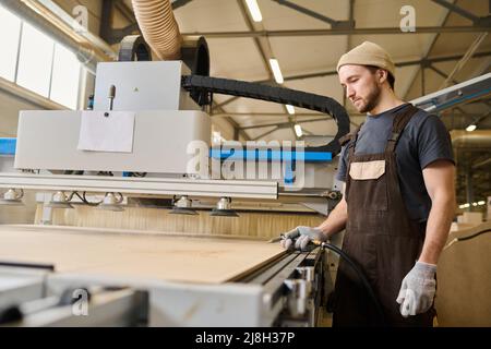 Junger Zimmermann in Schürze, der mit Holzbrettern an der modernen Maschine in der Möbelfabrik arbeitet Stockfoto