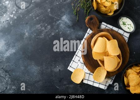 Ungesunde Lebensmittel. Snacks. Alle klassischen Kartoffelsnacks mit Erdnüssen, Popcorn- und Zwiebelringen und gesalzenen Brezeln in Schüsseln auf schwarzem Beton-Backgroun Stockfoto