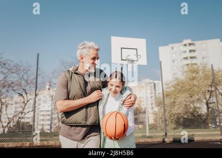 Glücklicher Vater und Tochter im Teenageralter umarmen und betrachten die Kamera draußen am Basketballplatz. Stockfoto