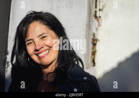 Porträt kaukasische Erwachsene Frau, lächelnd und Blick auf die Kamera, im Freien stehend in sonnenbeschienenen Innenhof. Stockfoto