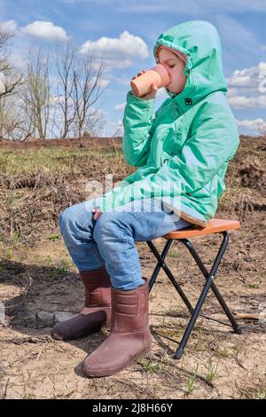 Ein positives glückliches kleines kaukasisches Mädchen in einer grünen Jacke sitzt auf einem Klappstuhl und trinkt bei einem Picknick im Frühjahr Wasser aus einem Glas. Stockfoto