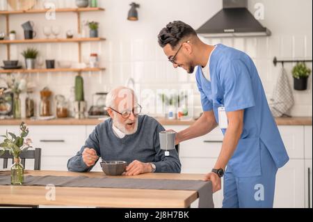 Fröhlicher Betreuer, der einem älteren Mann Tee serviert Stockfoto