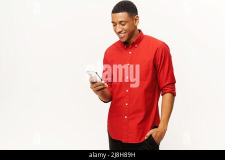 Lächelnder, optimistischer afroamerikanischer junger Mann in rotem Casual-Shirt mit Smartphone isoliert auf weißem, multirassischem Freiberufler oder Student, der online chattet, SMS, Messaging, Surfen im Internet, Kopierbereich Stockfoto