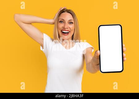 Froh schockiert emotionale hübsche junge europäische Dame in weißem T-Shirt zeigen Telefon mit leerem Bildschirm Stockfoto