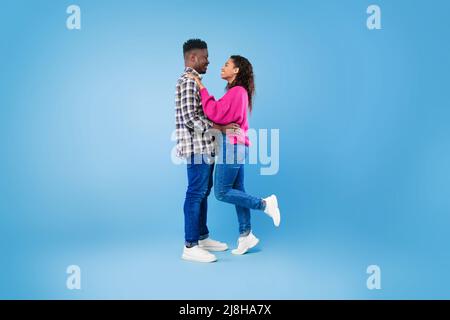 Seitenansicht eines glücklichen jungen afroamerikanischen Paares in legerer Kleidung, das sich umarmt und auf blauem Hintergrund anschaut Stockfoto