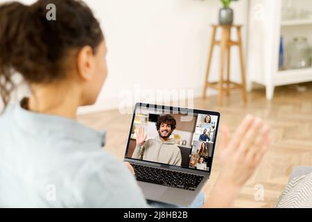 Nahaufnahme einer lockigen Frau, die auf dem Laptop-Bildschirm mit Personenprofilen winkt, mit einer Anwendung für die Fernkommunikation mit Kollegen, Webinar-Teilnehmern und Online-Treffen bei Pandemie Stockfoto