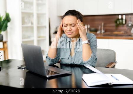 Frustrierte junge Frau, die einen Laptop benutzt, der Kopfschmerzen hat, im Heimbüro sitzt und den Kopf mit den Händen hält, sich krank und unwohl fühlt, sich ausbrennend und überfordert fühlt Stockfoto