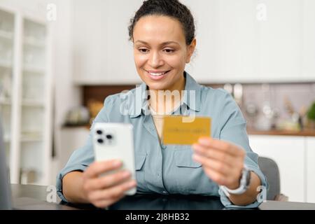 Eine fröhliche Frau, die die Smartphone-Shopping-App zum Online-Kauf nutzt, ihre Kreditkarte hält und Bankdaten eingibt, sitzt in entspannter Atmosphäre im Wohnzimmer zu Hause auf dem Sofa Stockfoto