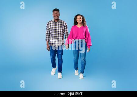 Die ganze Länge des tausendjährigen afroamerikanischen Paares, das die Hände hält und die Kamera auf blauem Studiohintergrund anlächelt Stockfoto