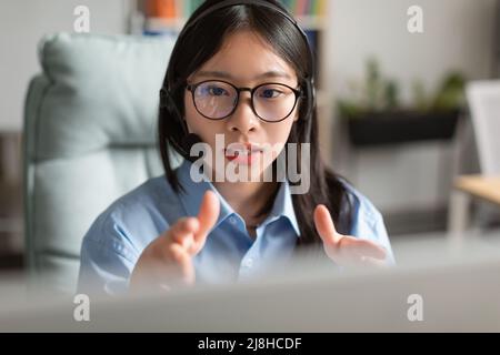 Asiatische Frau, die einen Laptop mit einem Headset am Schreibtisch trägt Stockfoto