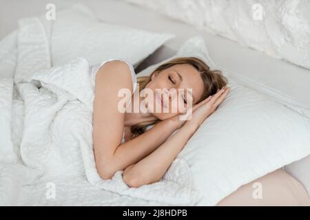 Fröhliche, hübsche junge blonde europäische Frau, die auf einem weichen Bett im weißen Schlafzimmer schläft Stockfoto