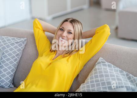 Fröhliche hübsche junge blonde europäische Frau in gelben Kleidern sitzt auf dem Sofa mit den Händen hinter dem Kopf Stockfoto