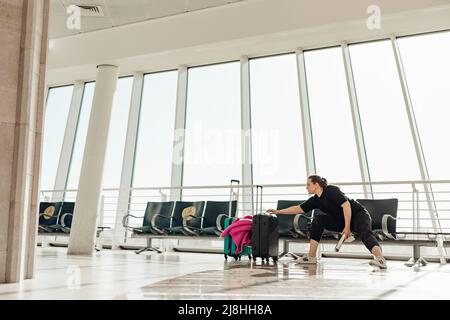 Passagier sitzt auf einem Stuhl in einem modernen hellen Flughafen mit großen Panoramafenstern in der Nähe von Koffern mit Kleidung und einem wartenden Flugzeug. Jung Stockfoto