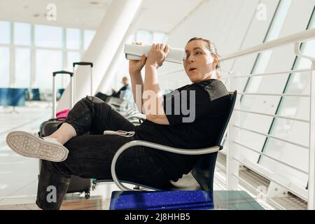 Passagier sitzt auf einem Stuhl in einem modernen hellen Flughafen in der Nähe von Koffern mit Kleidung, schaut durch die Röhre aus dem Magazin und hat Spaß Seitenansicht. Jung Stockfoto