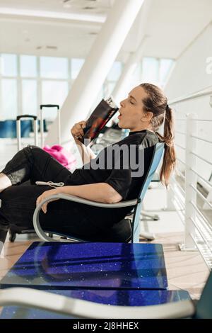 Junge Frau, die auf einem Stuhl auf dem Flughafen in der Nähe von Koffern mit Kleidung sitzt, unter Hitze leidet, gähnt und winkt, während sie auf das Flugzeug wartet Stockfoto