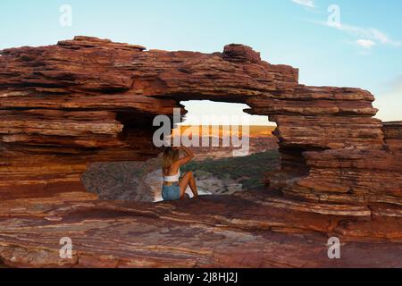 Porträt einer Frau in legerer Kleidung am natürlichen roten Felsenfenster mit spektakulärer Aussicht im Kalbarri-Nationalpark in Australien Stockfoto