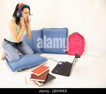 Gestresste junge Studentin, die online mit Kopfhörern, Büchern, Webkamera, Computer und Rucksack studiert Stockfoto