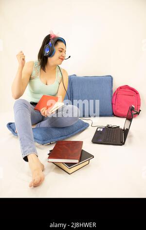 Glückliche junge Studentin, die mit Kopfhörern, Büchern, Webkamera, Computer und Rucksack studiert Stockfoto