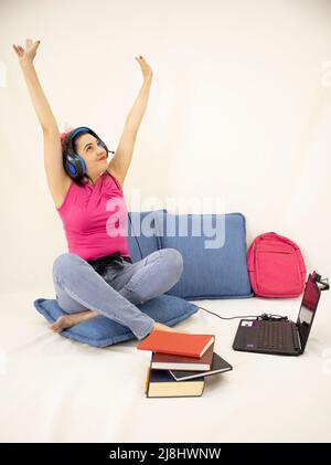 Glückliche junge Studentin, die mit Kopfhörern, Büchern, Webkamera, Computer und Rucksack studiert Stockfoto