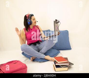 Junge Studentin, die in Online-Kursen winkt und mit Kopfhörern, Büchern, Webkamera, Computer und Rucksack studiert Stockfoto