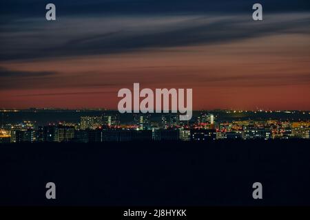 Nächtliche Stadtlandschaft mit Lichtern in den Stadthäusern. Mehrstöckige Gebäude nach Sonnenuntergang mit rotem Himmel am Abend Stockfoto