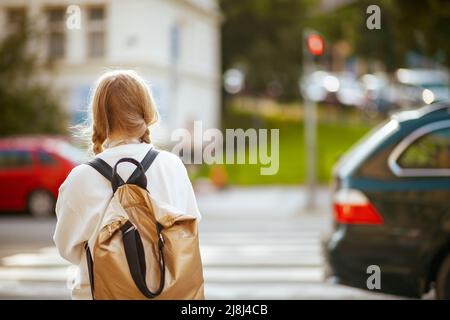 Von hinten gesehen trendiges Schulmädchen im weißen Sweatshirt mit Rucksack, der über den Crosswalk geht und in der Stadt draußen zur Schule geht. Stockfoto