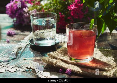 Rustikaler ästhetischer Outdoor-Tisch mit Glas Wasser, Kompott und schönen Blumen und Spitze Dekoration. Ästhetisches Picknick mit hellem Frühling, Sommerton Stockfoto