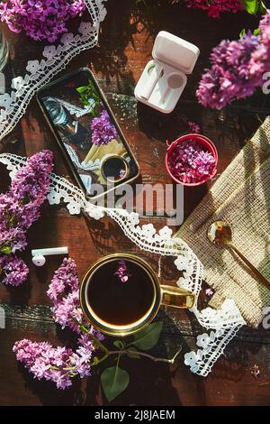 Rustikale ästhetische Outdoor-Tisch mit Glas Wasser, Tasse Kaffee, schöne Blumen und Spitze. Telefonbildschirm, kabellose Kopfhörer. Ästhetisches Picknick. St Stockfoto