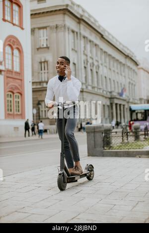 Hübscher junger Afroamerikaner mit Mobiltelefon, während er mit einem Elektroroller auf einer Straße steht Stockfoto