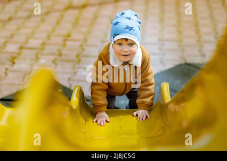 Fröhlicher lustiger kleiner Junge in einem blauen Hut und warmer Kleidung spielt auf dem Spielplatz und reitet von einer hellen bunten Rutsche in einem Kindergarten, in einem Stockfoto