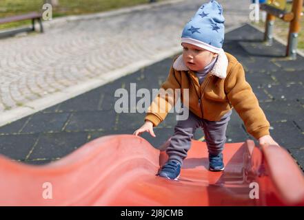 Fröhlicher lustiger kleiner Junge in einem blauen Hut und warmer Kleidung spielt auf dem Spielplatz und reitet von einer hellen bunten Rutsche in einem Kindergarten, in einem Stockfoto