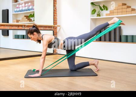 Sportliche Frau macht Power Yoga-Kick-Übungen mit Widerstandsbändern oder Riemen in voller Länge Seitenansicht in einem Fitnessstudio Stockfoto