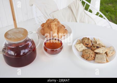 Sonntagsfrühstück im Hinterhof der Hütte. Frische süße Croissants mit Keksen auf dem Tisch. Transparente Teekane aus Glas mit gebrühtem Tee. Hochwertige Fotos Stockfoto