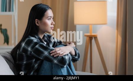 Seitenansicht traurige asiatische Frau, die zu Hause auf der Couch sitzt, sich unsicher über harte Entscheidung fühlt unglückliches schlechtes Gefühl geistige Probleme sitzt auf dem Sofa. Gedrückt Stockfoto