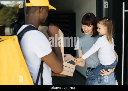 Schöne junge Frau, die ihre Tochter in den Armen hält, öffnet die Türen ihres Hauses und trifft auf einen Lieferer, der ihren Pappkarton mit Pizza aus dem Restaurant gibt. Stockfoto