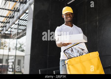 Afroamerikanischer Mann Kurier von Express-Lieferung, Online-Shopping-Konzept. Delivery Mann trägt gelben Thermorucksack und Karton, während wai Stockfoto