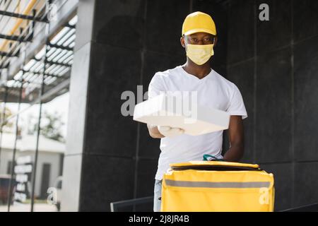 Delivery Mann trägt gelben thermischen Rucksack und Karton, während der Bestellung für den Käufer geben, trägt Gesichtsschutzmaske, während covid-19 Pandemie arbeiten. Stockfoto