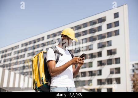 Attraktiver afroamerikanischer Lebensmittellieferungskurier, der vor der Kamera auf der Straße posiert. Lächelnder Mann mit einer gelben Mütze und einem wärmeisolierten Lebensmittelbeutel auf dem Rücken mit einem Telefon in den Händen. Stockfoto